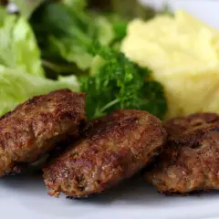 Drei goldbraune bayerische Fleischpflanzerl liegen auf einem Teller neben cremigem Kartoffelpüree und frischem Salat.