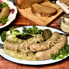 Saure Bratwurst mit Zwiebeln und Petersilie auf einem Teller, dazu Brot und ein Glas Senf.