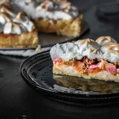 Ein Stück Rhabarberkuchen mit Baiser, das auf einem schwarzen Teller liegt. Der Kuchen hat eine goldbraune Kruste und eine saftige Rhabarberfüllung.