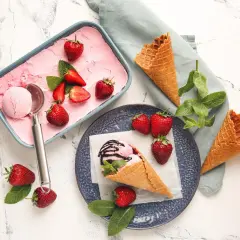 Erdbeer-Eis in einer Schale, garniert mit frischen Erdbeeren und Minze. Daneben leere Waffeln und eine Eiskugel im Hörnchen.