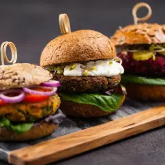 Drei appetitliche vegetarische Gerichte: Burger mit bunten Patties, frischem Gemüse und cremigen Saucen auf rustikalem Holzbrett.