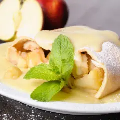 Ein köstlicher Apfelstrudel mit Eiermilch, bestäubt mit Puderzucker und garniert mit Minze. Daneben liegen Äpfel.