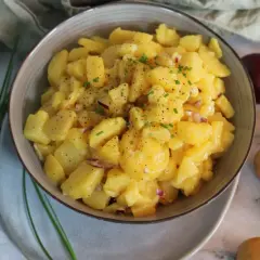 Schüssel mit leckerem Kartoffelsalat ohne Mayo, garniert mit Schnittlauch und Pfeffer. Daneben liegen frische Kartoffeln und eine rote Zwiebel.