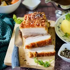 Ein saftiger Schweinebraten mit knuspriger Kruste, auf einem Holzbrett angeschnitten, daneben Klöße und Soße.