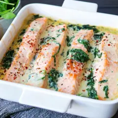 Vier saftige Lachsfilets in cremiger Spinatsauce, bestreut mit Gewürzen, in einer weißen Auflaufform. Daneben liegen frischer Spinat und eine Zitronenspalte.