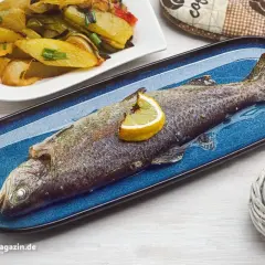 Eine ganze Forelle im Backofen, garniert mit einer Zitronenspalte, liegt auf einem blauen Teller. Daneben Kartoffelspalten.