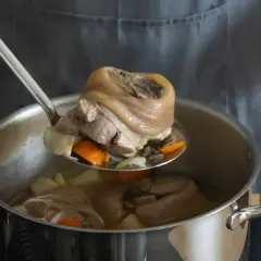 Eisbein zubereiten im Topf: Ein Koch hebt mit einer Schöpfkelle ein gekochtes Eisbein aus einem Topf mit Gemüse und Brühe.