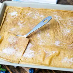 Großer, goldbrauner Pfannkuchen vom Blech, bestreut mit Zucker und Zimt. Ein Stück wird mit einem Löffel herausgehoben. Blaubeeren und Minze garnieren.