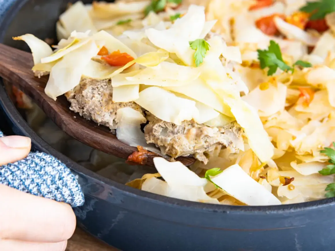 Ein Holzlöffel hebt eine Portion Krauttopf mit Hackfleisch aus einem Topf. Das Gericht ist im Ofen gebacken und mit Petersilie garniert.