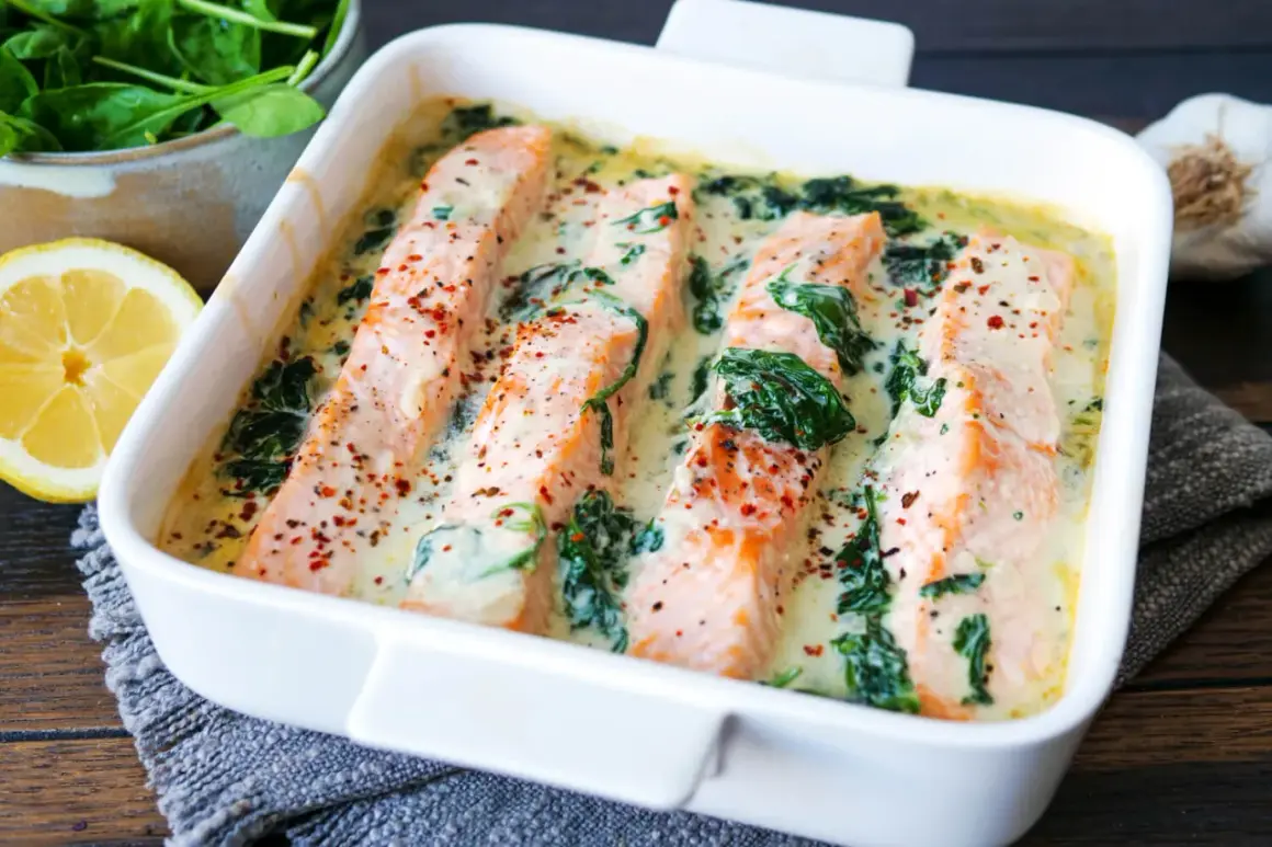 Vier saftige Lachsfilets in cremiger Spinatsauce, bestreut mit Gewürzen, in einer weißen Auflaufform. Daneben liegen frischer Spinat und eine Zitronenspalte.