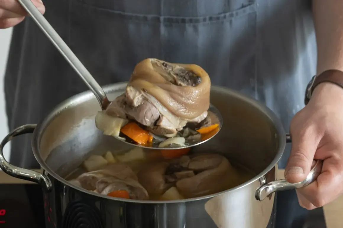 Eisbein zubereiten im Topf: Ein Koch hebt mit einer Schöpfkelle ein gekochtes Eisbein aus einem Topf mit Gemüse und Brühe.