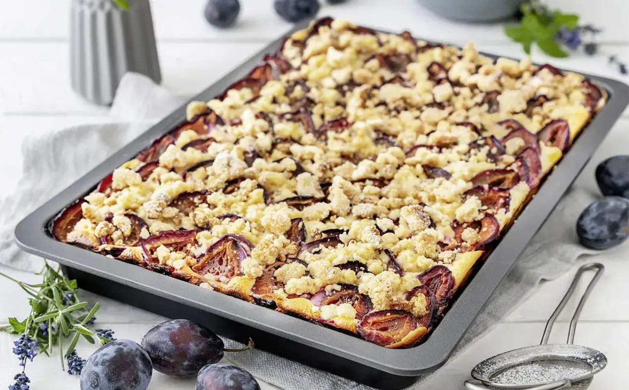 Ein frisch gebackener, gesunder pflaumenkuchen mit Streuseln in einer Backform. Frische Pflaumen und Kräuter liegen daneben.