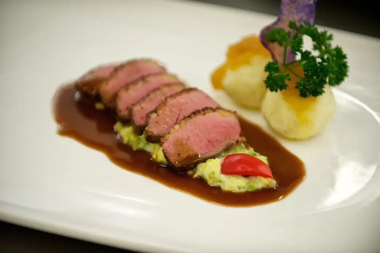 Zartrosa gebratene entenbrust mit wirsing, Kartoffelklößen und einer dunklen Sauce auf einem weißen Teller.