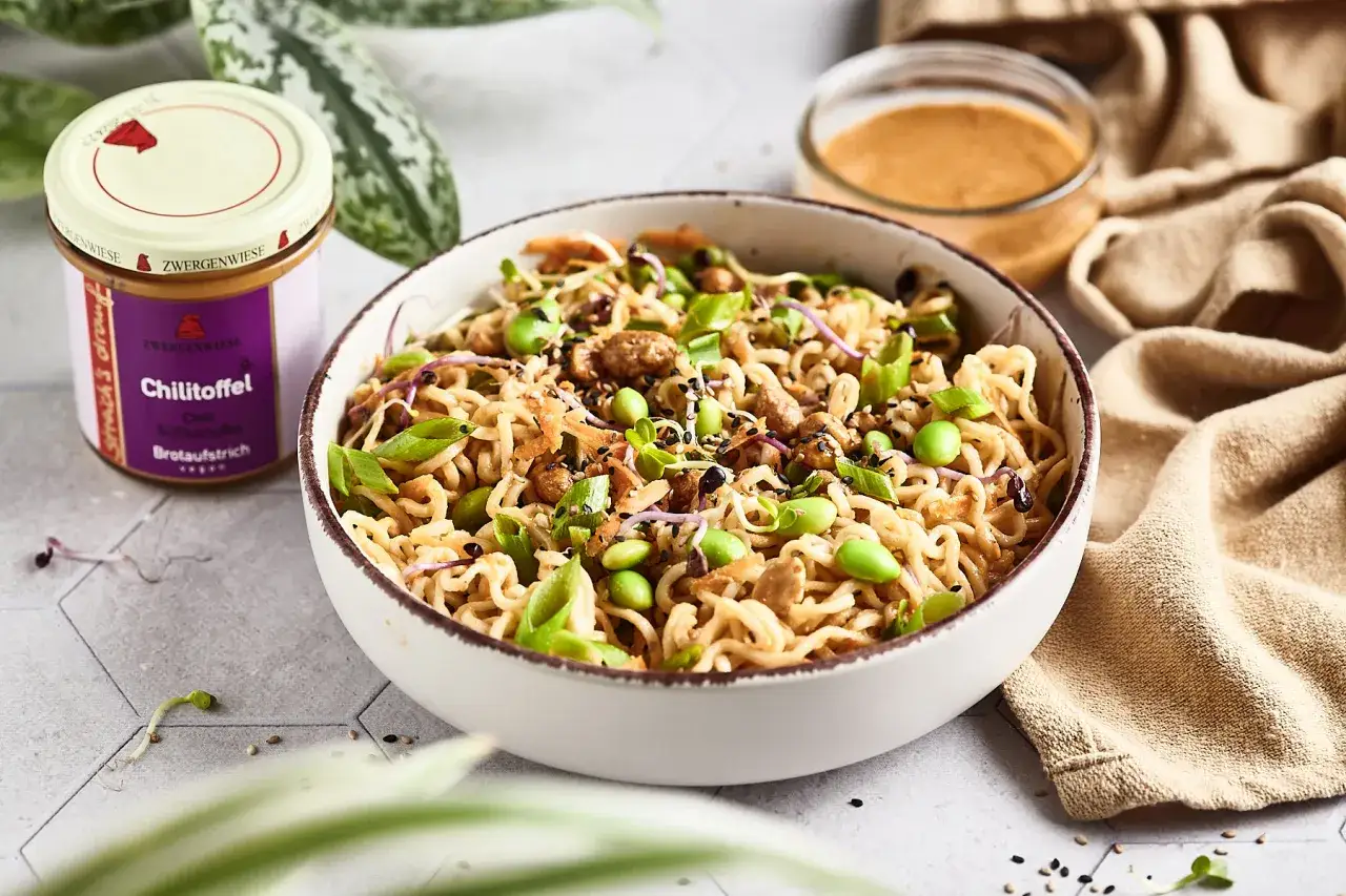 Herzhafter mie nudelsalat mit Edamame, Kresse und gerösteten Zwiebeln. Ein Glas Chili-Aufstrich steht daneben.