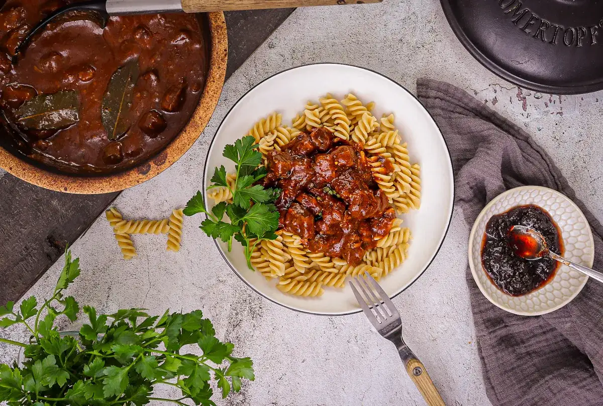 Herzhaftes Rindergulasch Rezept mit Fusilli-Nudeln, garniert mit Petersilie. Dazu eine kleine Schale mit dunkler Beerenmarmelade.