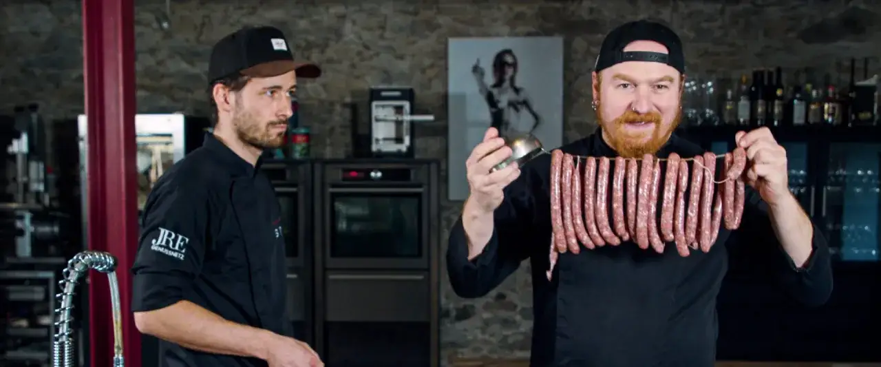 Zwei Köche präsentieren eine Reihe von rohen Wildbratwürsten, die an einer Schnur hängen. Das beste Wildbratwurst Rezept wird hier enthüllt!