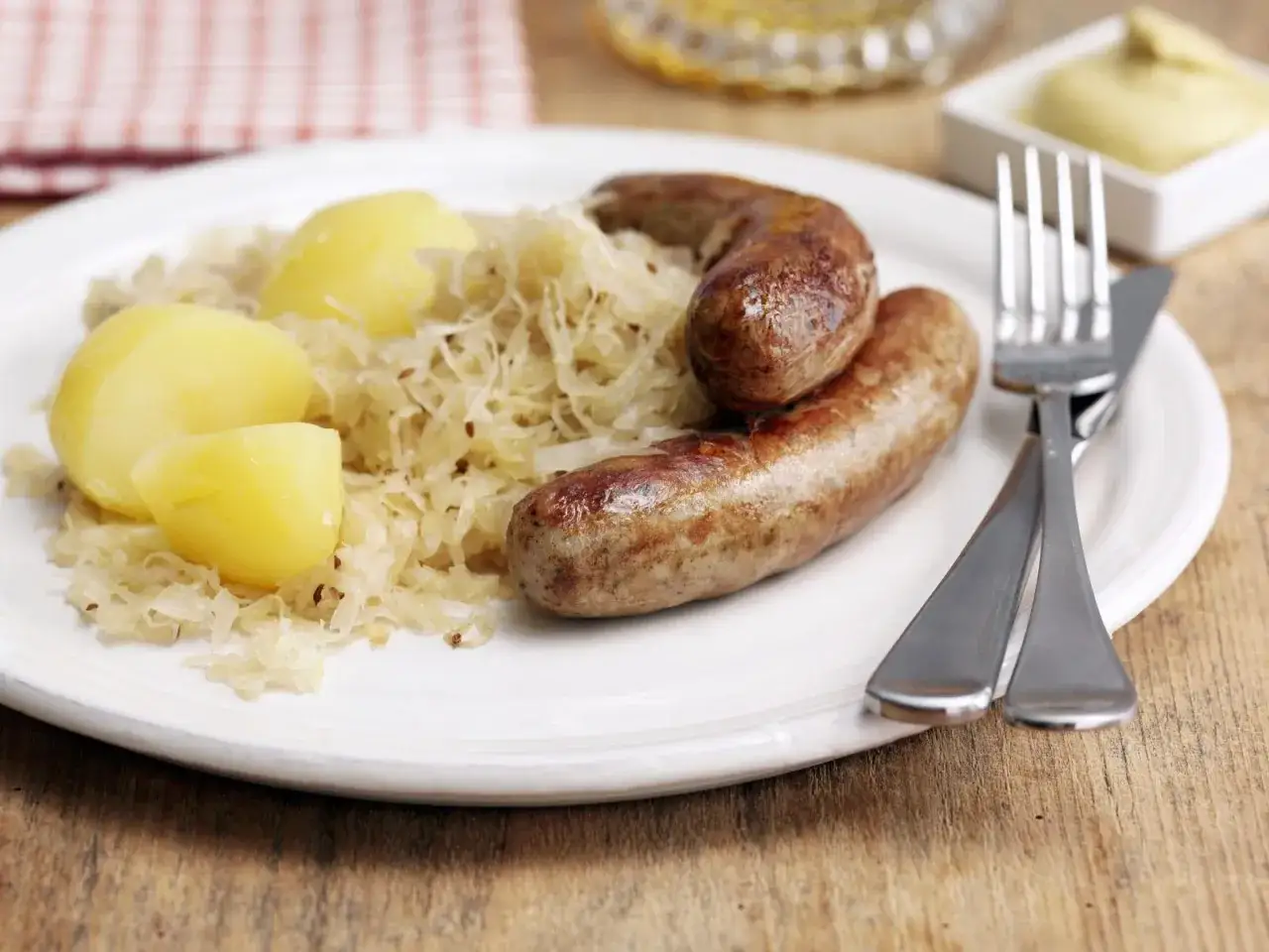 Zwei Bratwürste vom Vortag liegen auf Sauerkraut mit Kartoffeln. Daneben liegen Messer und Gabel.