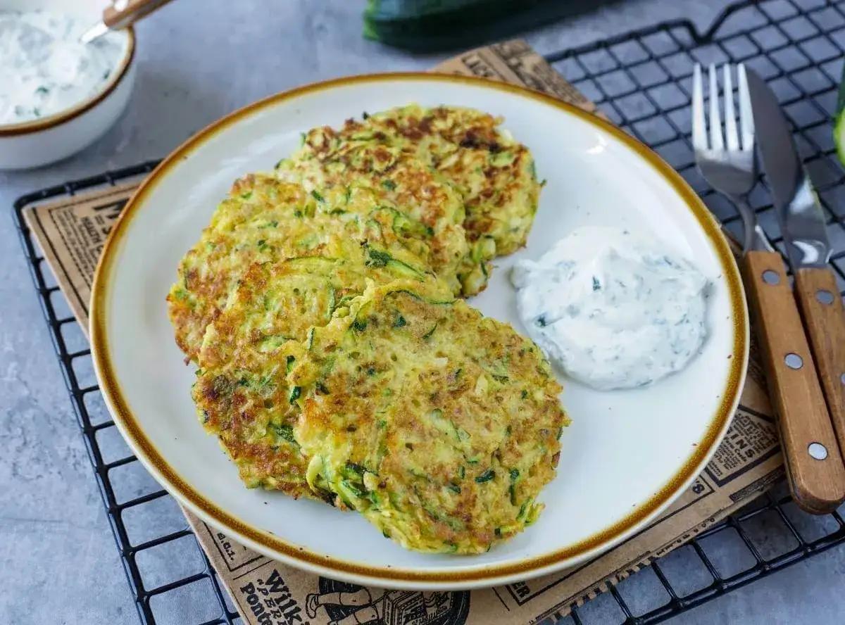 Goldbraune Zucchini-Puffer mit Kräuterquark auf einem Teller, daneben ein Messer und eine Gabel.