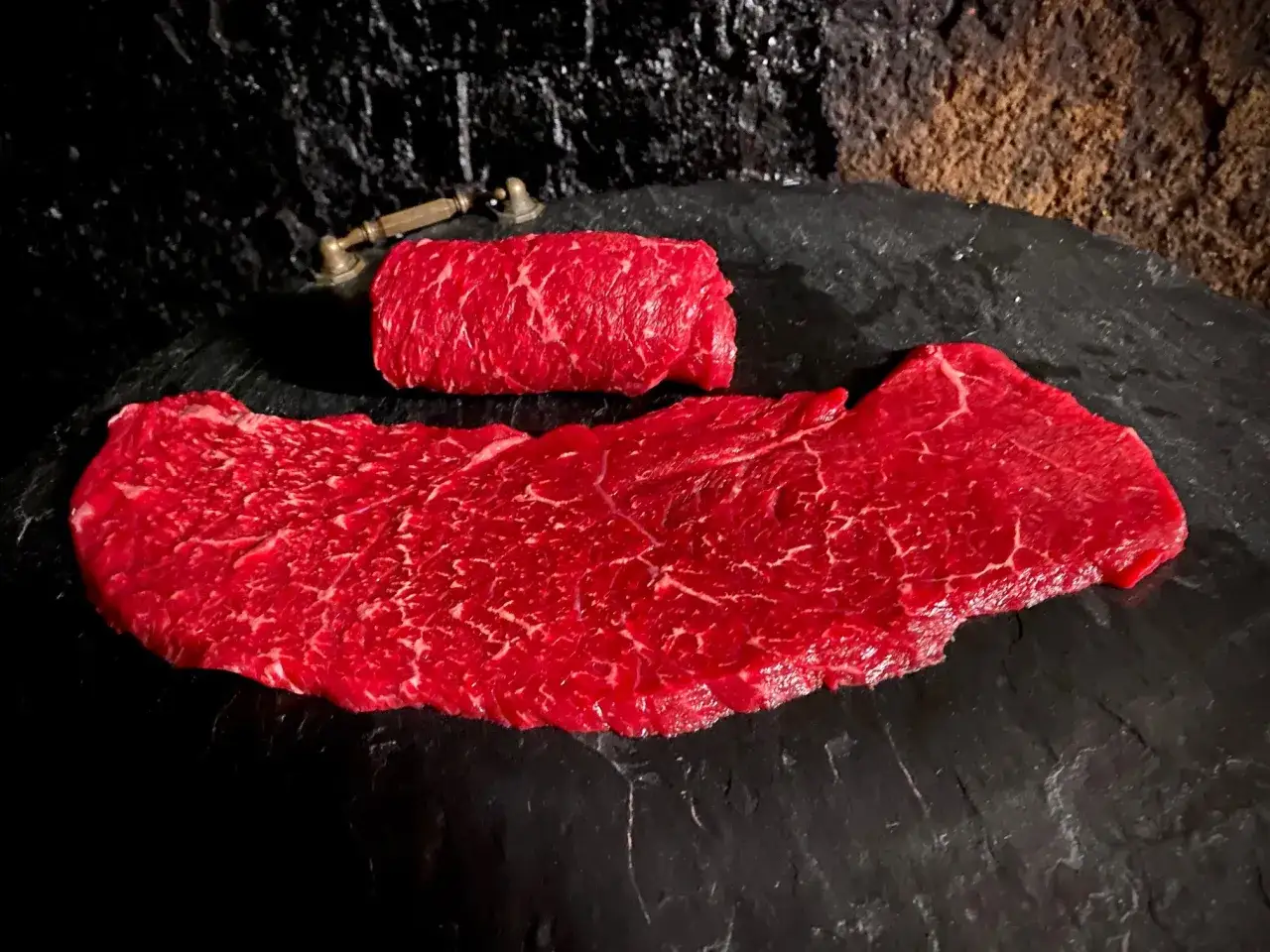 Rohe Schweinerouladen, welches Fleisch eignet sich am besten? Zarte, marmorierte Scheiben liegen auf dunklem Stein.