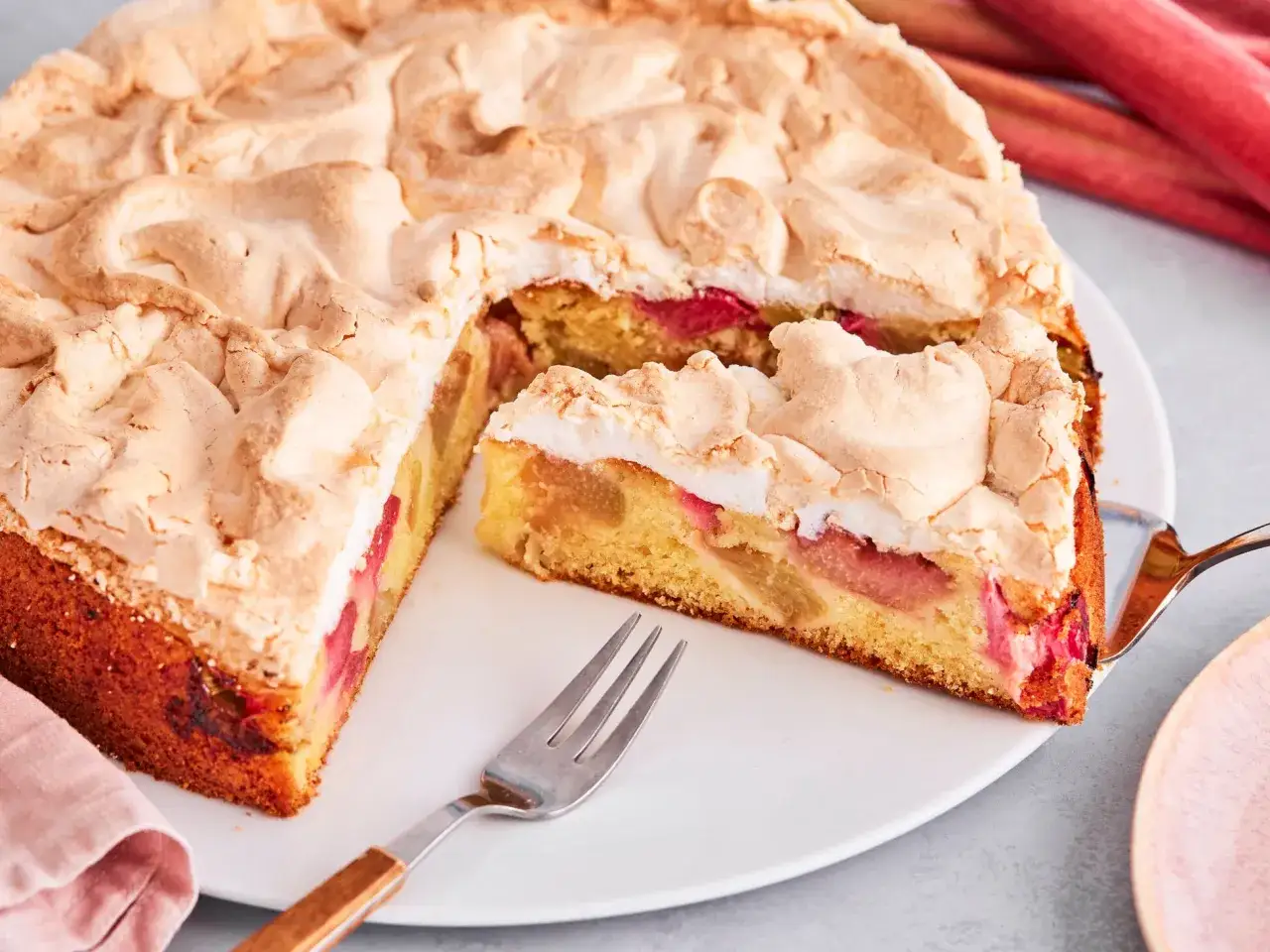 Ein saftiger Rhabarberkuchen mit luftigem Baiser, bereit zum Genießen. Daneben liegen frische Rhabarberstangen.