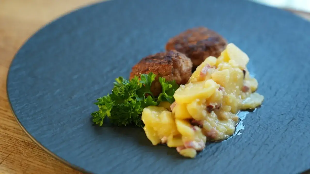 Zwei goldbraune vegetarische Frikadellen mit Kartoffelsalat und Petersilie auf einem dunklen Teller.