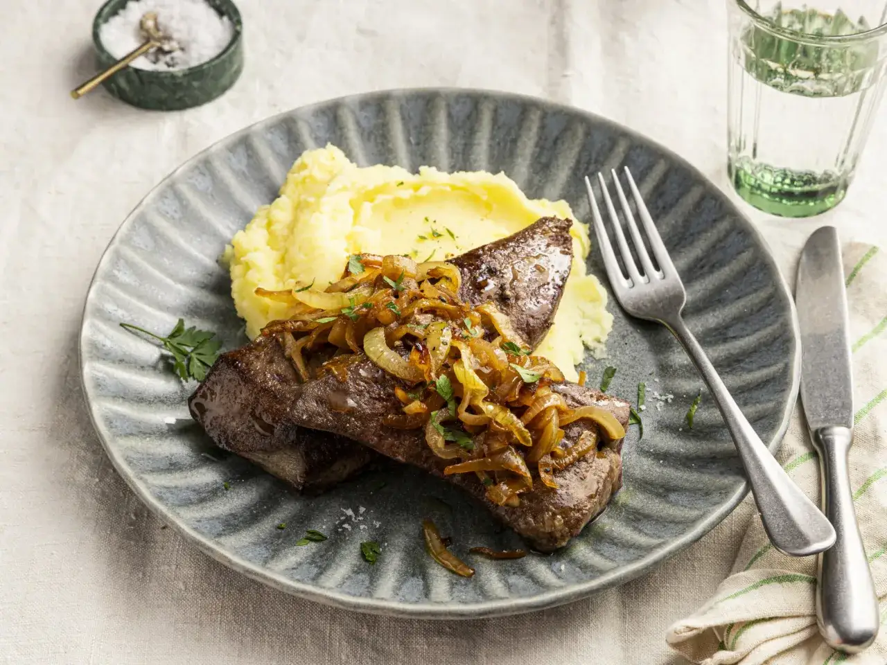 Leber braten ohne Mehl: Zarte Leber mit karamellisierten Zwiebeln und cremigem Kartoffelpüree auf einem grauen Teller.