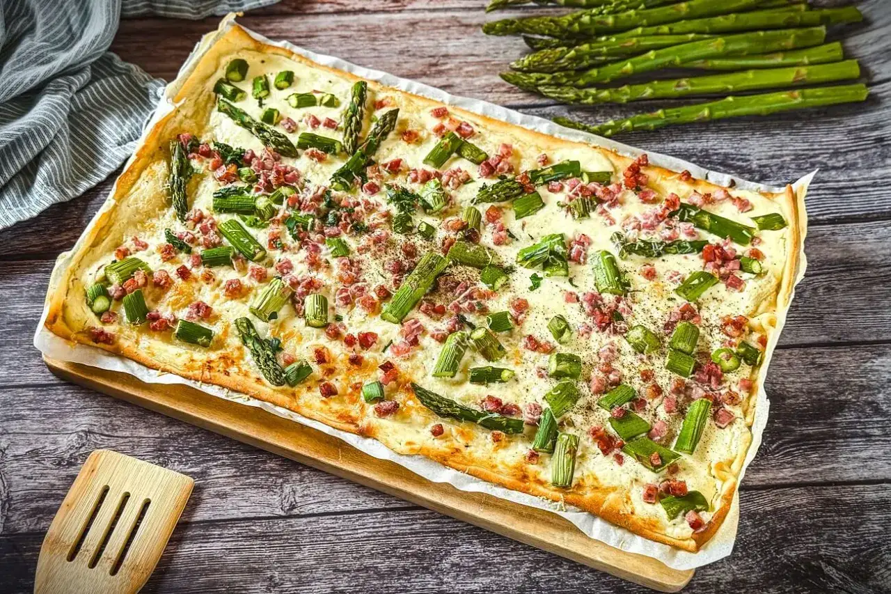Ein rechteckiger Spargel Flammkuchen mit Speck und einer cremigen Käsesauce, garniert mit frischem Spargel und Kräutern.