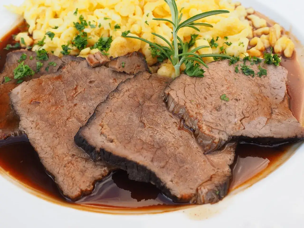 Saftige Scheiben Sauerbraten in dunkler Sauce, serviert mit Spätzle und garniert mit Rosmarin. Die ideale Sauerbraten Temperatur für perfekten Genuss.
