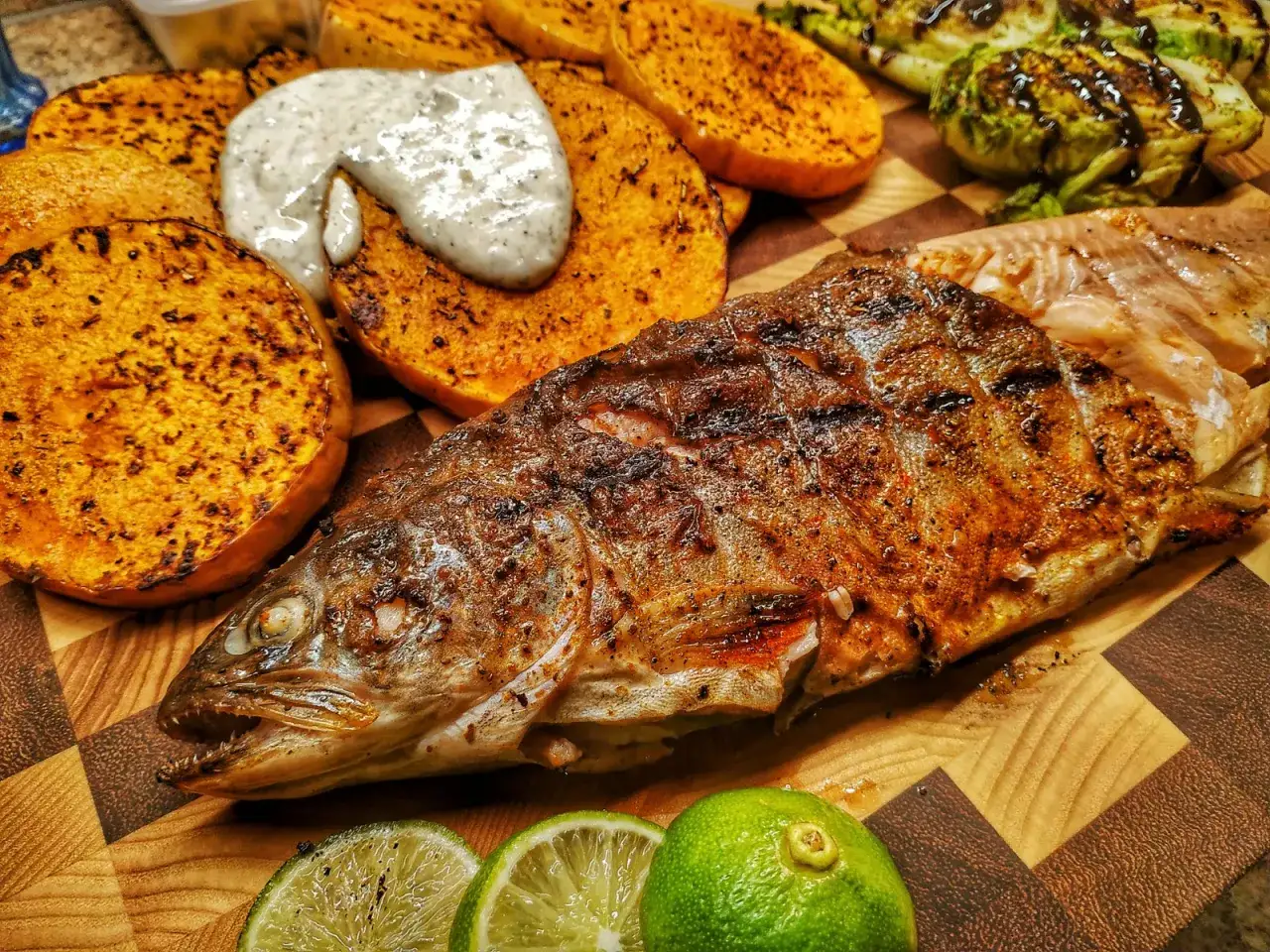 Saibling grillen: Ein ganzer, gegrillter Fisch liegt neben gerösteten Süßkartoffelscheiben und einem Dip. Dazu gibt es Limettenspalten.
