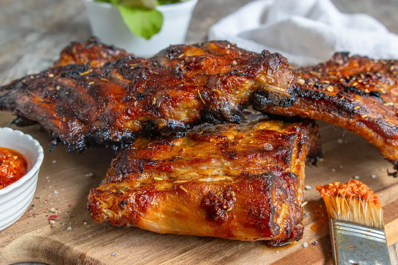 Saftige spareribs im backofen, glasiert und mit Gewürzen bestreut, liegen auf einem Holzbrett. Daneben eine Schale mit scharfer Sauce und ein Pinsel.