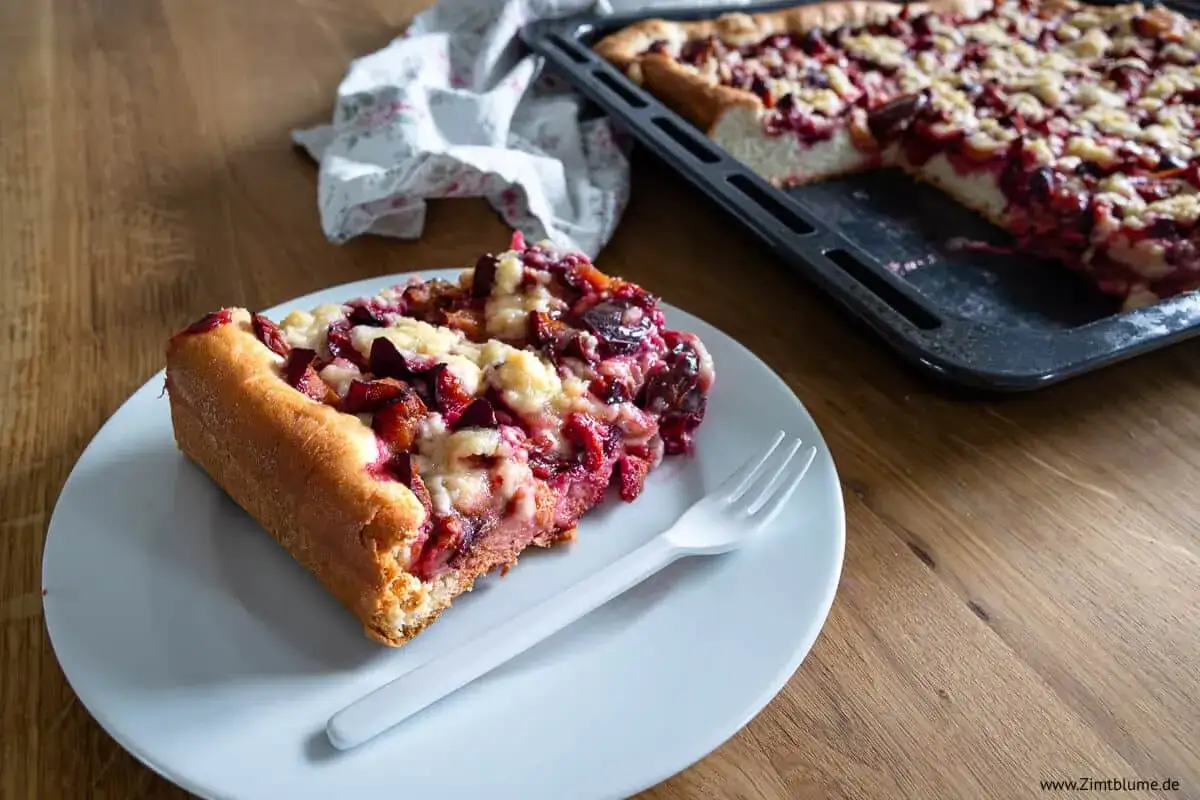 Ein Stück saftiger Zwetschgenkuchen auf Hefeteig mit Streuseln, daneben der ganze Kuchen im Blech.